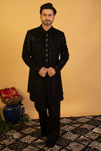 A man wearing a black embellished sherwani jacket with a matching black shirt and trousers, standing against a warm beige background.