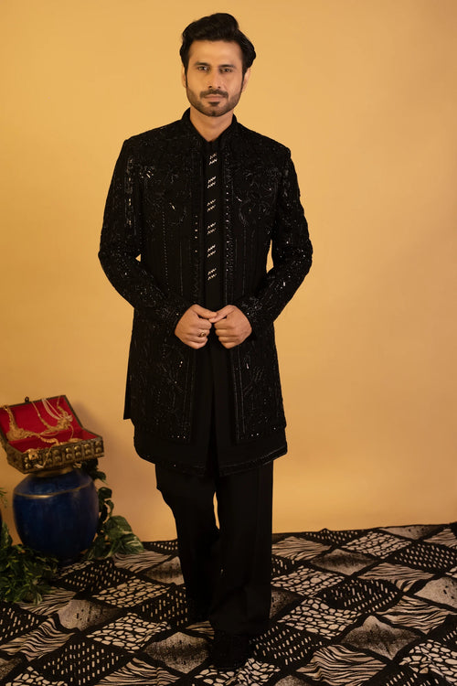 A man wearing a black embellished sherwani jacket with a matching black shirt and trousers, standing against a warm beige background.